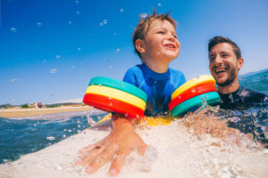 Photo of a cheerful little boy trying to stand up on a surfboard with a little help from his dad holding him from behind