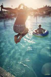 Little boy and his sister are playing in swimming pool. The boy is floating on water and the firl is jumping into the pool.